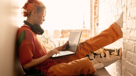 Seorang wanita muda dengan rambut oranye duduk nyaman di dekat jendela dengan headphone over-the-ear di lehernya melihat laptop Windows 11-nya.