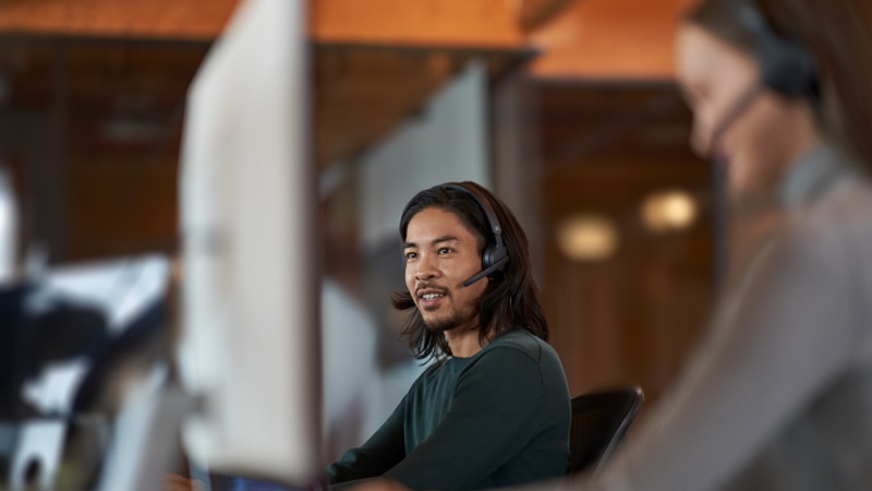 Homem com cabelo comprido usando um fone de ouvido, trabalhando em um computador em um escritório, com um colega em segundo plano